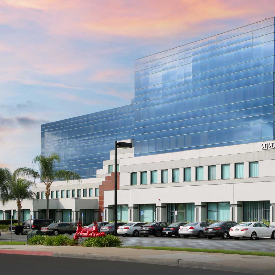 A modern office building with a reflective glass facade and a white lower section. The building is surrounded by palm trees and has a parking lot in front with several cars parked. The sky above is colorful with shades of pink, orange, and blue, indicating either sunrise or sunset.