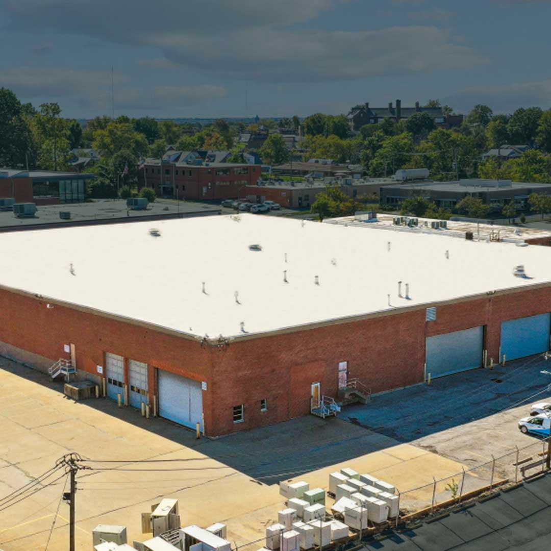 A large industrial building with a flat white roof and multiple loading docks with roll-up doors, surrounded by a paved area with pallets and equipment. Trees and other buildings are visible in the background.
