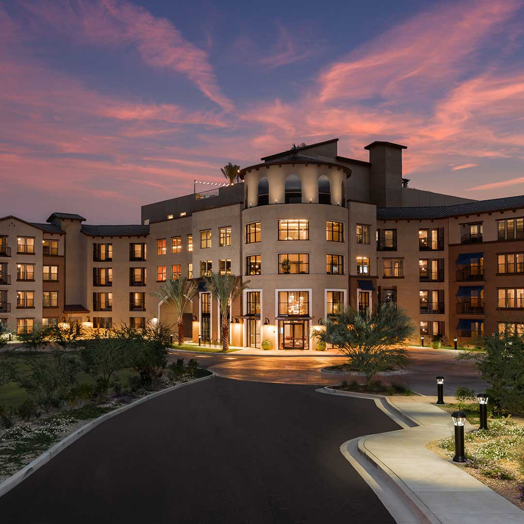 A large multi-story building, possibly a hotel or apartment complex, with many illuminated windows at sunset. The sky is painted in vibrant pink and orange hues, and the entrance is framed by streetlights and well-kept landscaping.