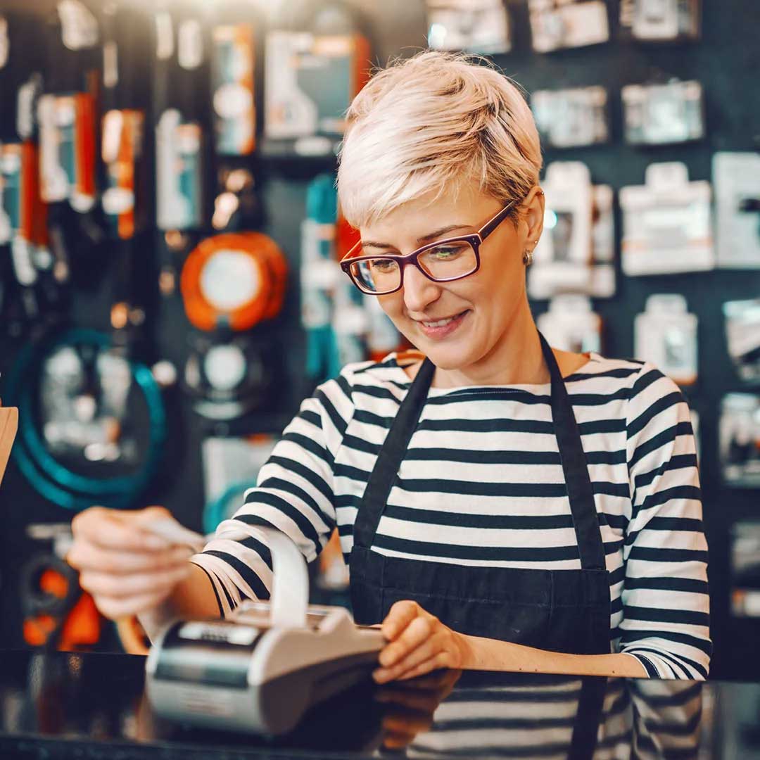 A female clerk in a striped shirt and apron works on a calculator or register.