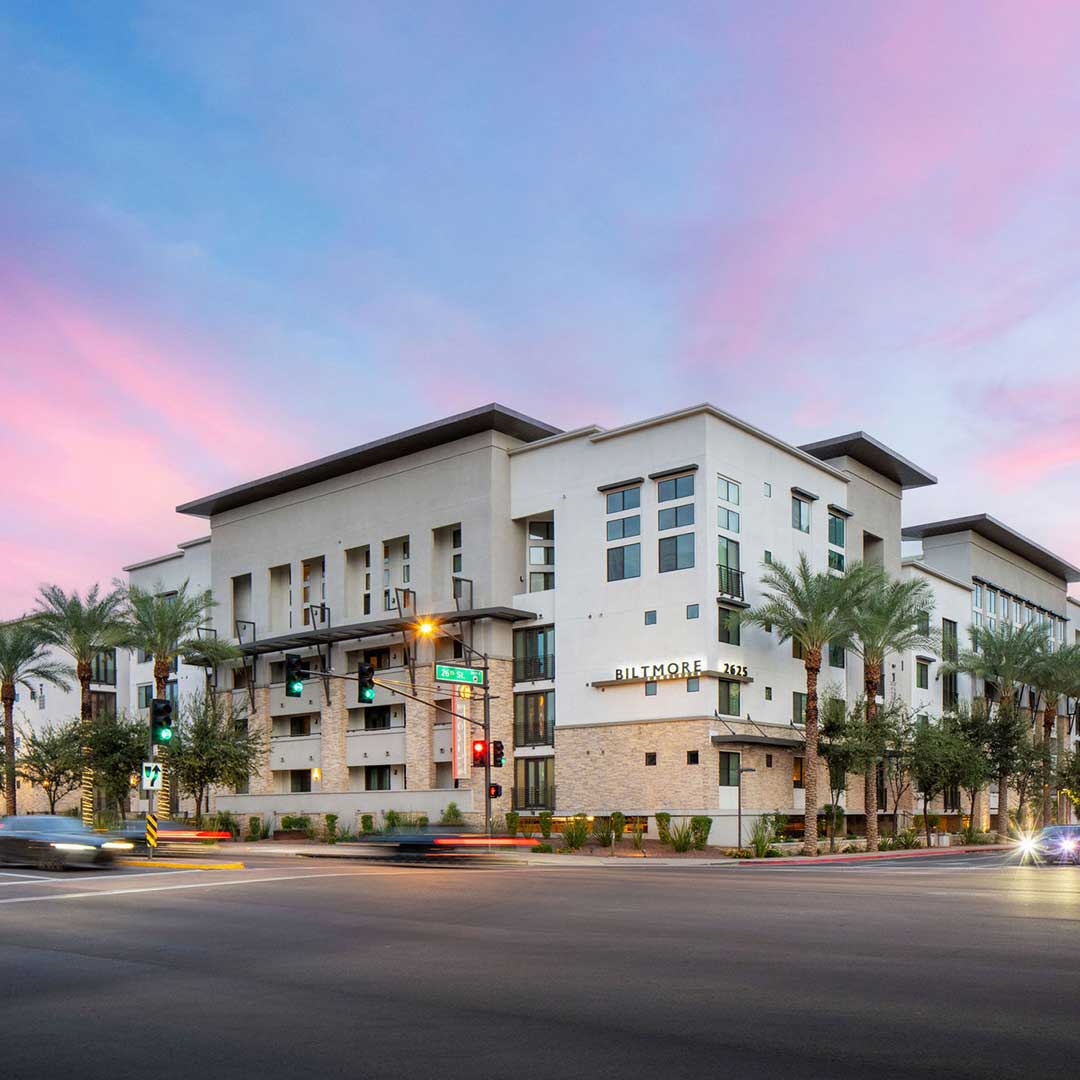 Modern white apartment building on a street corner at sunset, with palm trees and traffic lights.