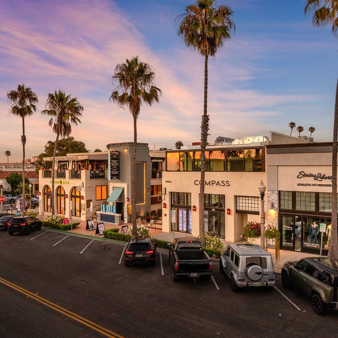 Street view of La Plaza in La Jolla with palm trees, shops, and parked cars at sunset.