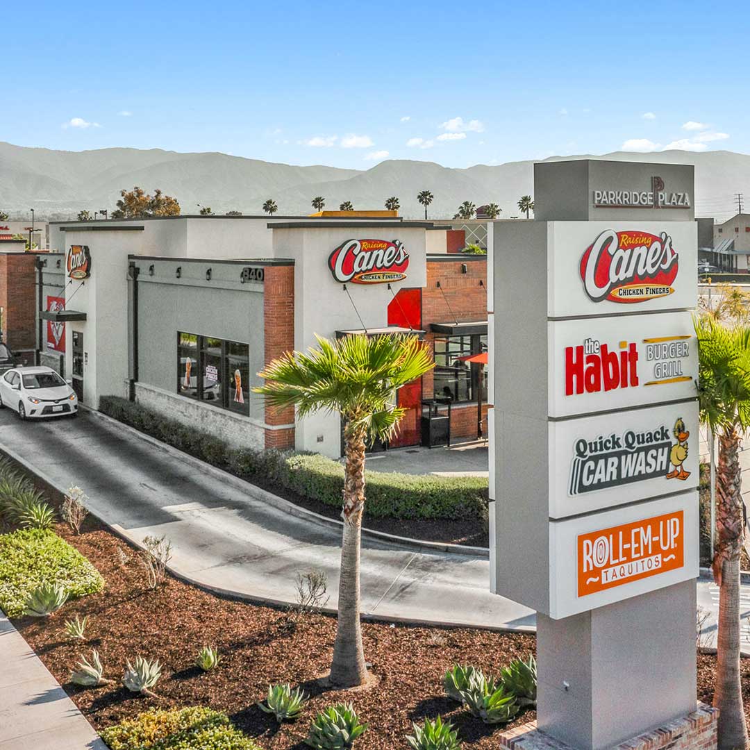Raising Cane’s restaurant with drive-thru and plaza sign listing The Habit, Quick Quack Car Wash, and Roll-Em-Up Taquitos