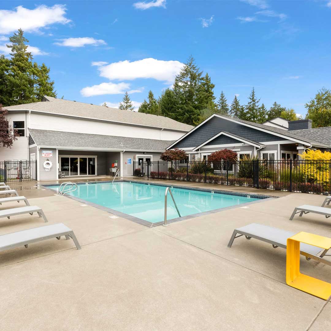 Outdoor pool with grey and blue clubhouse and white and yellow lounge chairs.