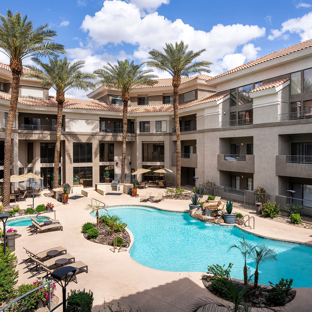 Courtyard pool area with lounge chairs, palm trees, and multi-story apartment building under blue sky.