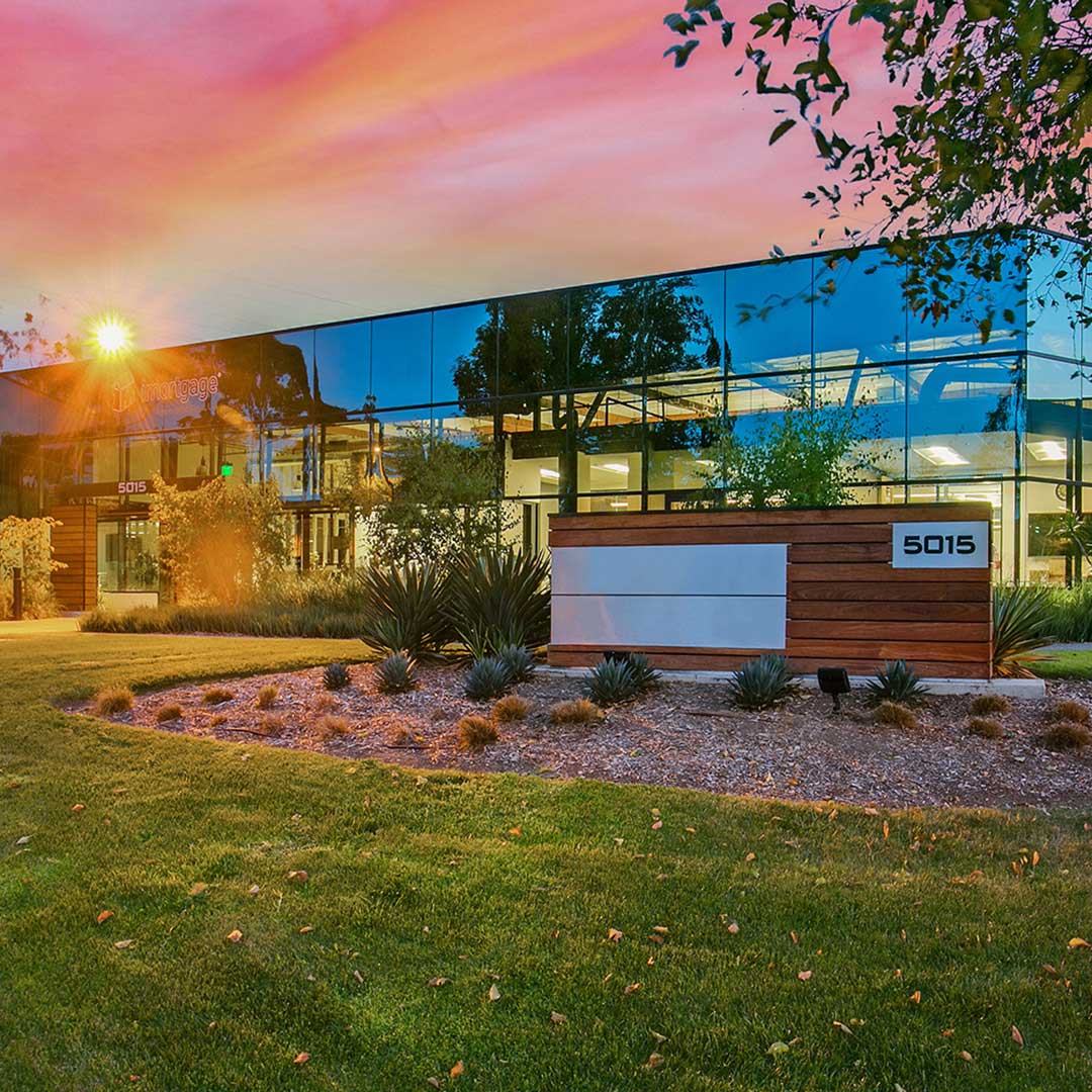 Modern office building with reflective glass facade at 5015 Shoreham Place, San Diego, surrounded by landscaped greenery and plants, under a vibrant pink and orange sky at sunrise or sunset.