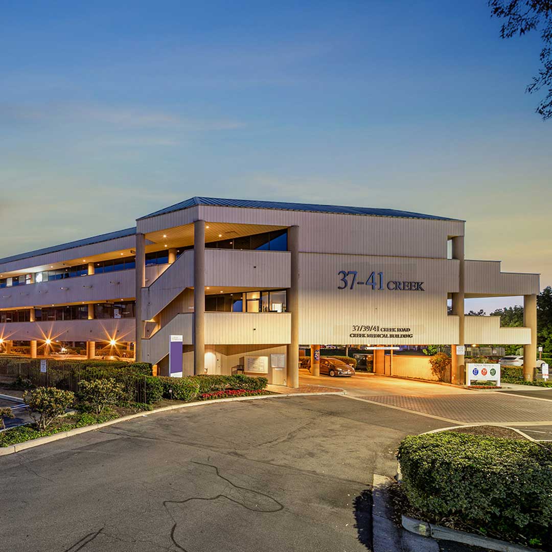 A three-story commercial building with the address '37-41 Creek' prominently displayed on the front. The building has a modern design with beige exterior walls and large windows. External staircases are visible on the left side. The entrance is well-lit, and a driveway leads to an underground parking area. The surrounding area features landscaped bushes and trees, with a clear sky at dusk in the background.