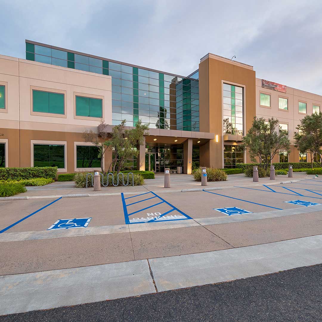 A modern office building with a glass facade and beige exterior walls. The building has three stories and features large windows. In front of the building, there are several handicap parking spaces marked with blue paint, as well as a bike rack and some landscaping with bushes and small trees. The entrance to the building is covered by an overhang supported by columns.