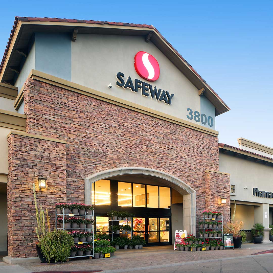 Exterior of a Safeway grocery store with a stone facade, arched entrance, and the Safeway logo above the doorway. The store address '3800' is visible, with potted plants and flowers displayed outside under a clear blue sky.