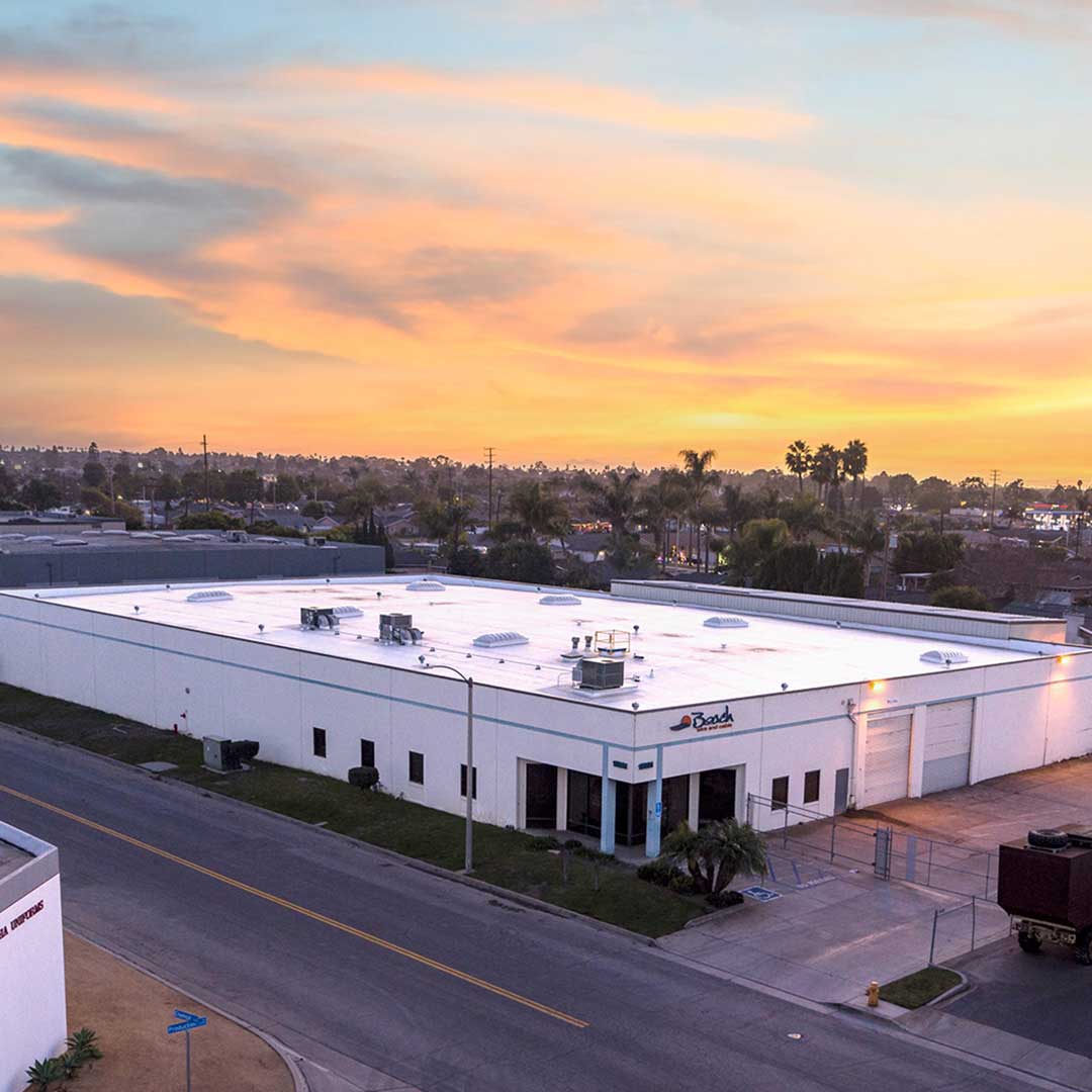 An aerial view of a large, white industrial building with a flat roof and several rooftop units. The building is situated on a corner lot with surrounding streets and greenery. The sky is filled with vibrant colors from the setting sun, creating a picturesque backdrop. There are palm trees and other buildings visible in the distance.