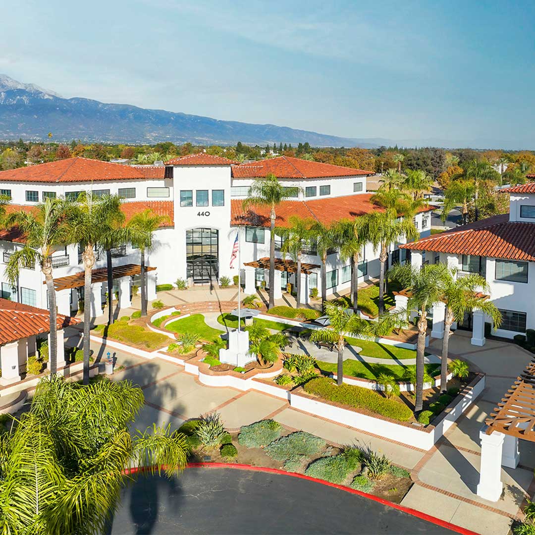 White office building with red tile roof, palm trees, and mountain backdrop on a sunny day.