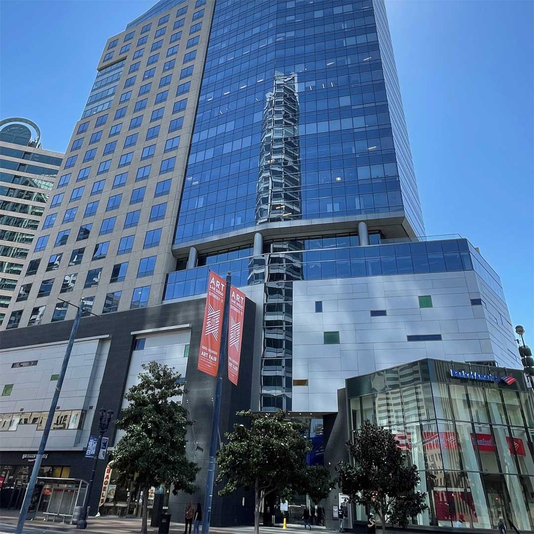 Modern office building with a glass facade reflecting nearby structures, featuring white and grey panels on the lower section and blue-tinted windows on the upper section. Two red banners promoting an art event hang in front. Trees, smaller buildings, and a Bank of America branch are visible at street level under a clear blue sky.