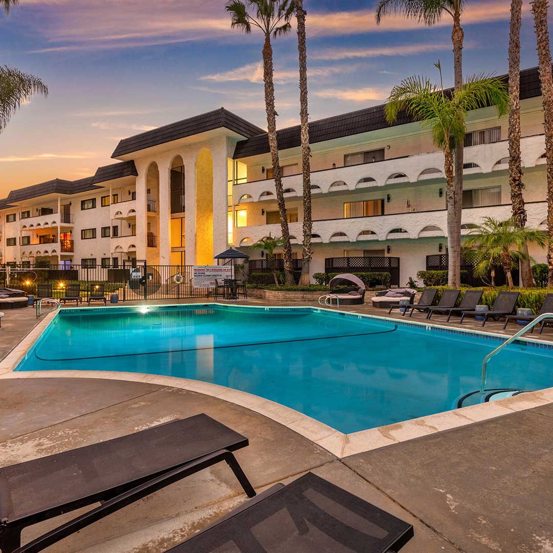 A serene outdoor swimming pool area at sunset, surrounded by lounge chairs and palm trees. In the background, there is a multi-story building with arched balconies and large columns. The sky is painted with warm hues of orange and pink, adding to the tranquil atmosphere.