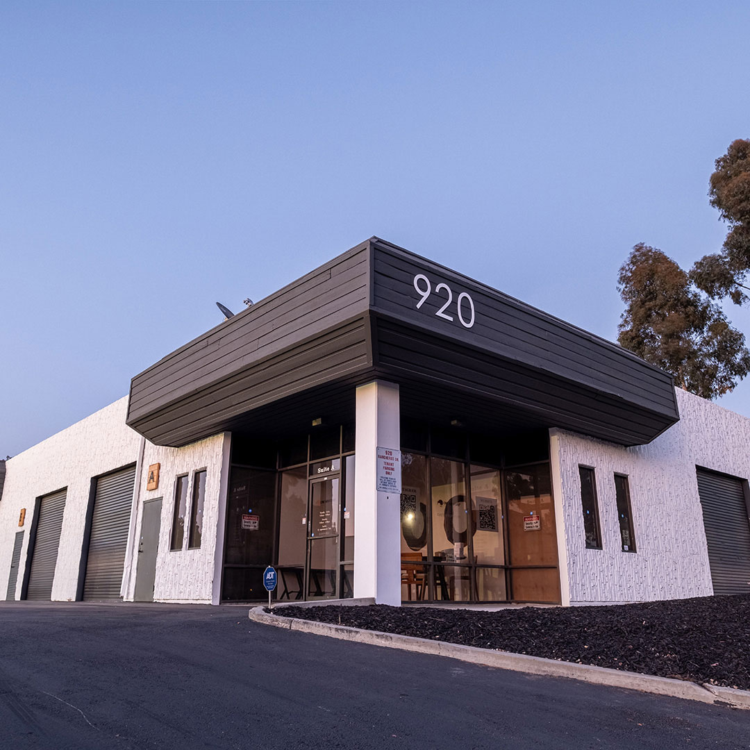 Modern commercial building with a white exterior and black accents, labeled '920' above the entrance. The structure features large glass windows and doors at the front, smaller windows and garage-style doors along the side, with a paved driveway and landscaped mulch area. Trees and a clear sky are visible in the background.