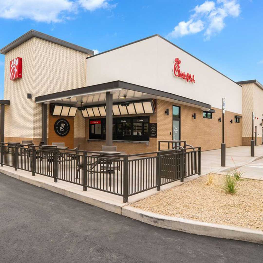 A Chick-fil-A restaurant with a modern beige brick exterior and white facade, featuring an outdoor seating area enclosed by black metal railings. The Chick-fil-A logo is prominently displayed on the building under clear blue skies.