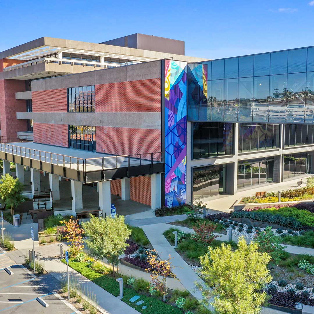A modern building with a combination of brick and glass exterior, featuring colorful murals on the corners. The building is surrounded by landscaped gardens with various plants and trees, as well as paved walkways. There is a parking lot adjacent to the building. The sky is clear and blue.