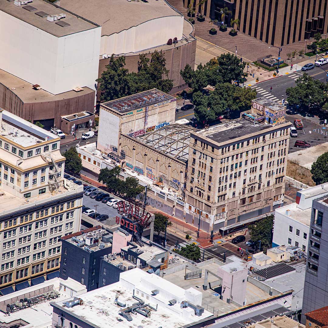 An aerial view of an urban area featuring several buildings. The central focus is on two large, older buildings with visible graffiti and signs of wear. One building has a partially collapsed roof structure. Surrounding these are other commercial and office buildings, streets with parked cars, and some greenery in the form of trees lining the streets. The image captures a mix of architectural styles amidst a bustling city environment.