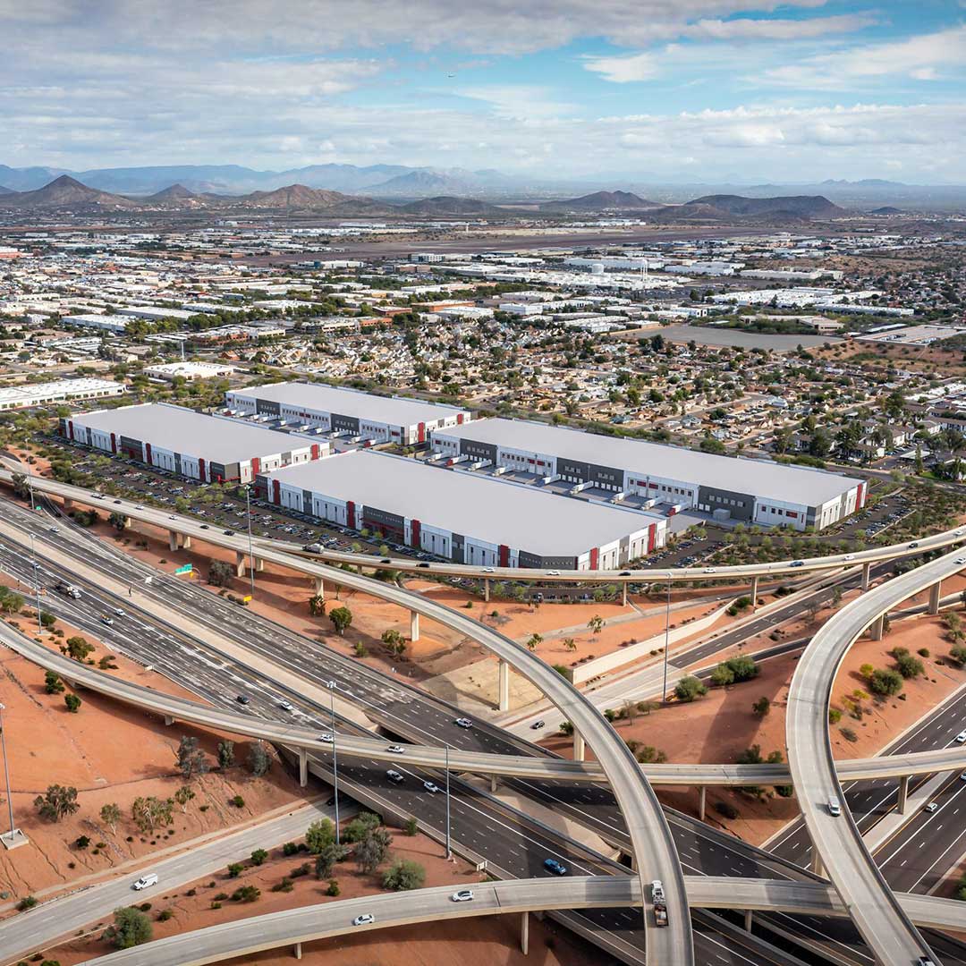 An aerial view of a large industrial area with multiple warehouse buildings. The warehouses have white and gray exteriors with red accents. Surrounding the warehouses are parking lots and roads. In the foreground, a complex network of highways and overpasses is visible with vehicles traveling on them. The background features a sprawling urban landscape with residential areas, commercial buildings, and distant mountains under a partly cloudy sky.