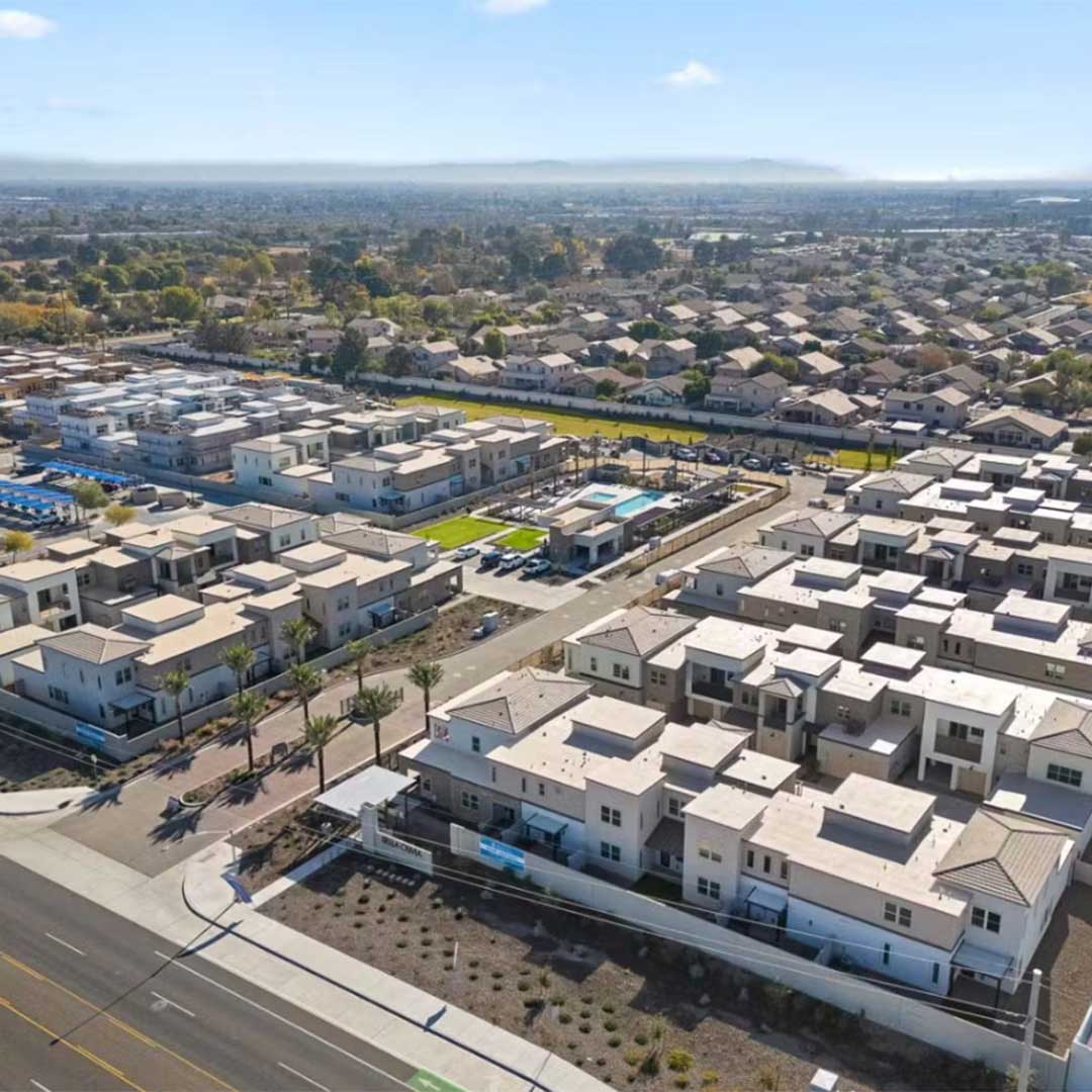 An aerial view of a residential neighborhood featuring modern, flat-roofed houses. The area is densely packed with homes, and there are some green spaces and a swimming pool visible in the center. The neighborhood is surrounded by more traditional suburban housing in the background, with a main road running along the bottom edge of the image. The sky is clear with a few clouds, and there are distant hills on the horizon.
