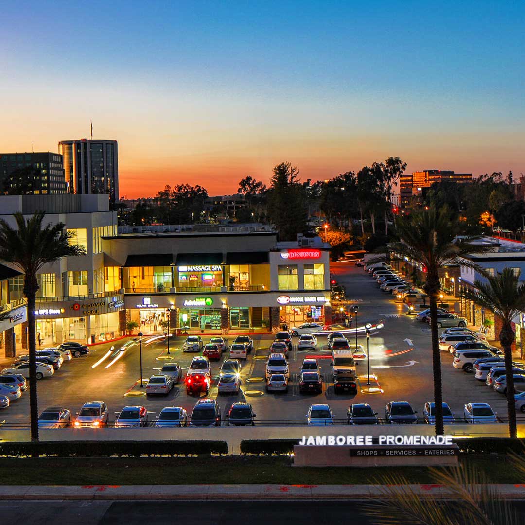 Jamboree Promenade shopping center at sunset with parked cars and illuminated storefronts.
