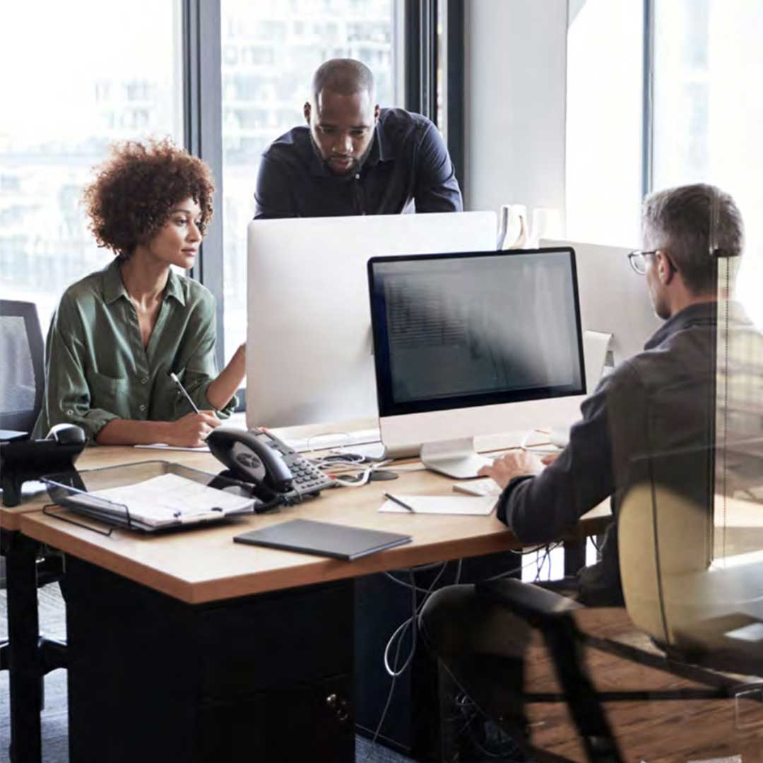 Three people are working together in an office setting. One person is seated at a desk, typing on a computer keyboard, while another stands behind the desk looking at the screen. A third person is seated next to the desk, writing on a notepad. The office features large windows with a cityscape view in the background. The desk holds a telephone, papers, and electronic devices.