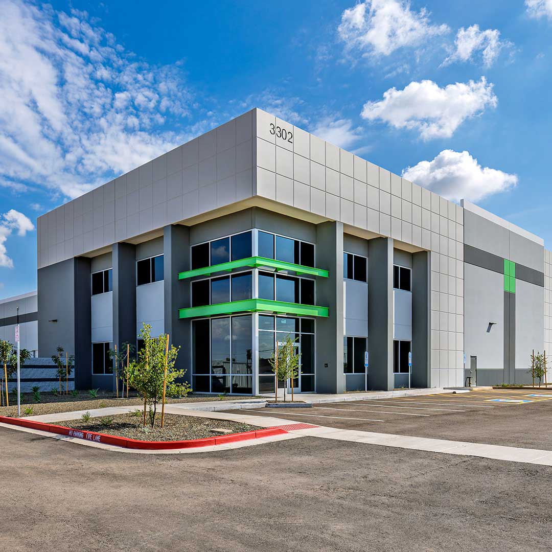 A modern commercial building with a grey and white exterior, featuring green accents around the windows. The building has large glass windows and is located in a parking lot with a few small trees planted around it. The address "3302" is visible on the top corner of the building. The sky is clear with some scattered clouds
