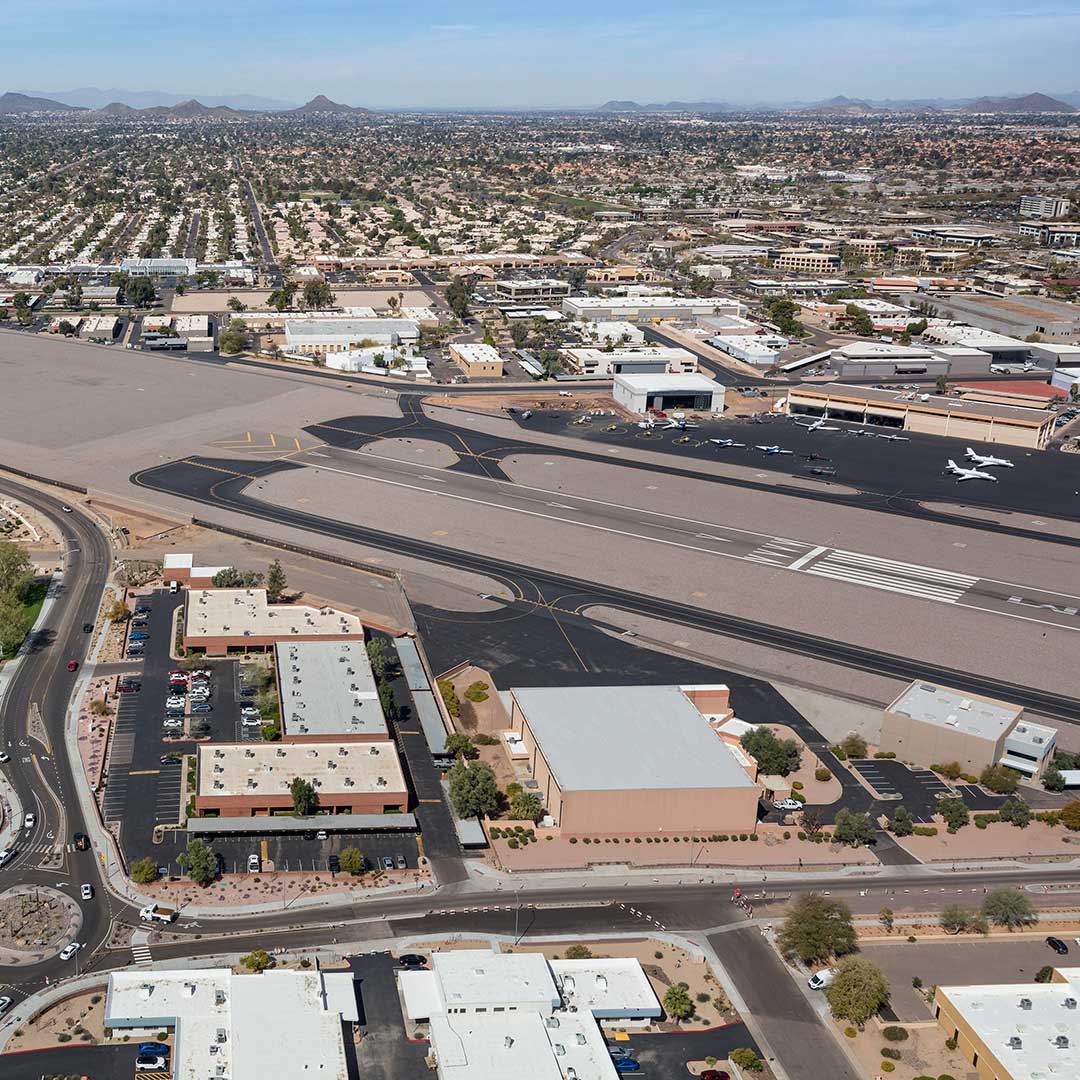 An aerial view of an airport with multiple runways and taxiways. Several buildings and hangars are visible around the airport, along with a few airplanes parked on the tarmac. The surrounding area includes a mix of commercial and residential buildings, with a grid-like pattern of streets extending into the distance. The landscape is flat, with mountains visible on the horizon under a clear blue sky.