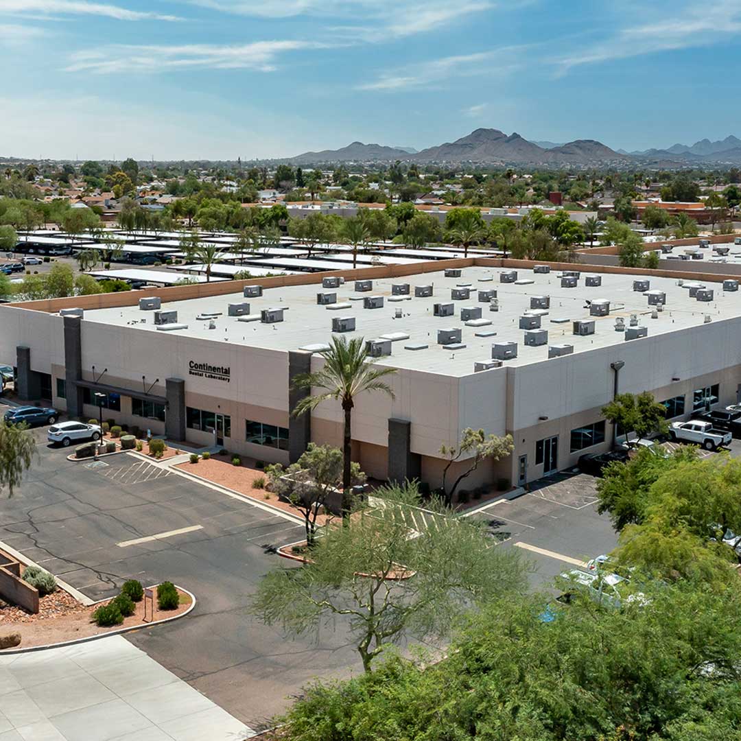Aerial view of a commercial building with the sign 'Continental' on the side. The building has a large flat roof with multiple HVAC units. The surrounding area includes parking lots, trees, and other buildings. In the background, there are mountains under a partly cloudy sky.
