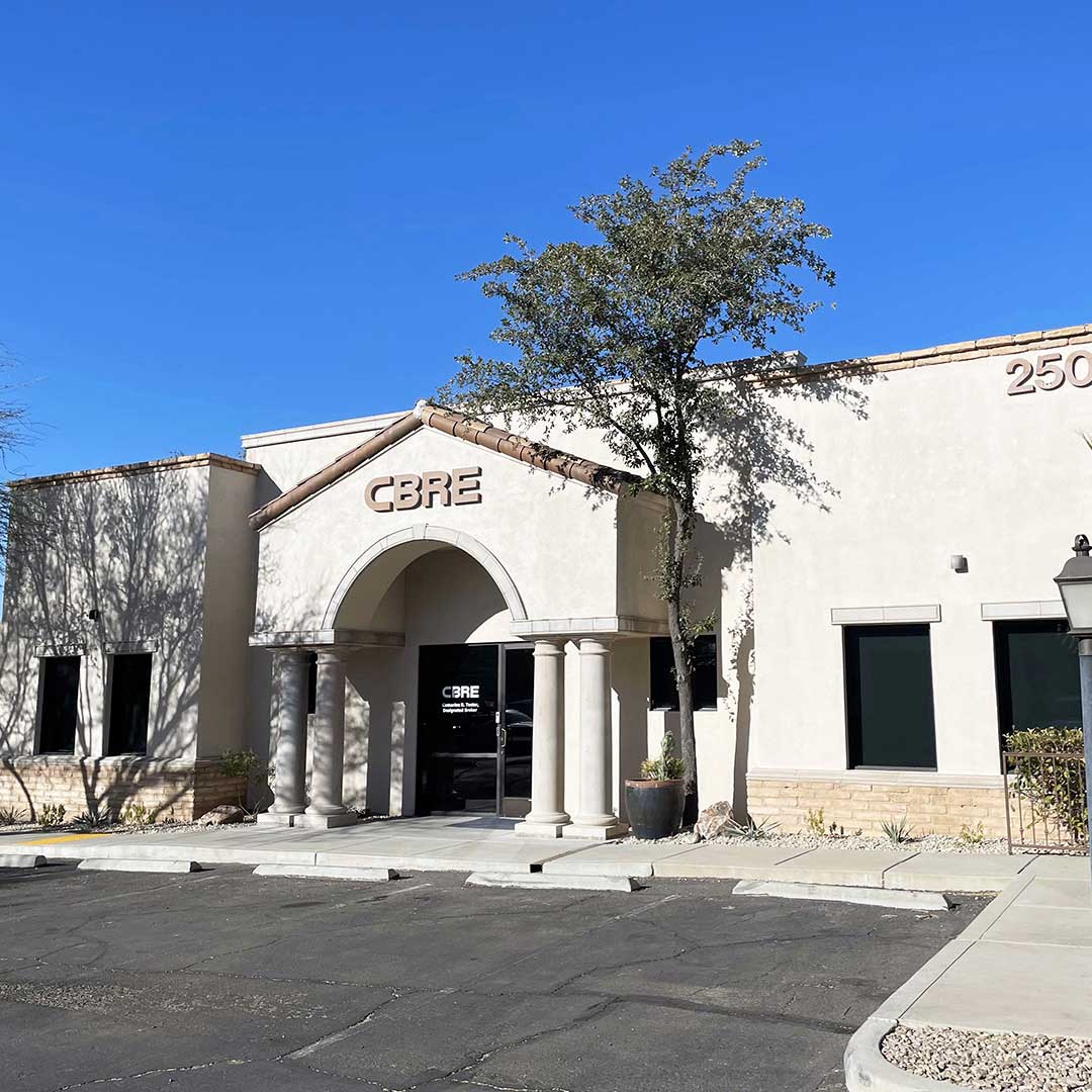 A single-story commercial building with a beige exterior and a flat roof. The entrance features an arched portico supported by four columns. The building has the letters "CBRE" above the entrance and the number "2502" on the right side of the facade. There are two large windows on either side of the entrance, and a small tree is planted near the front.
