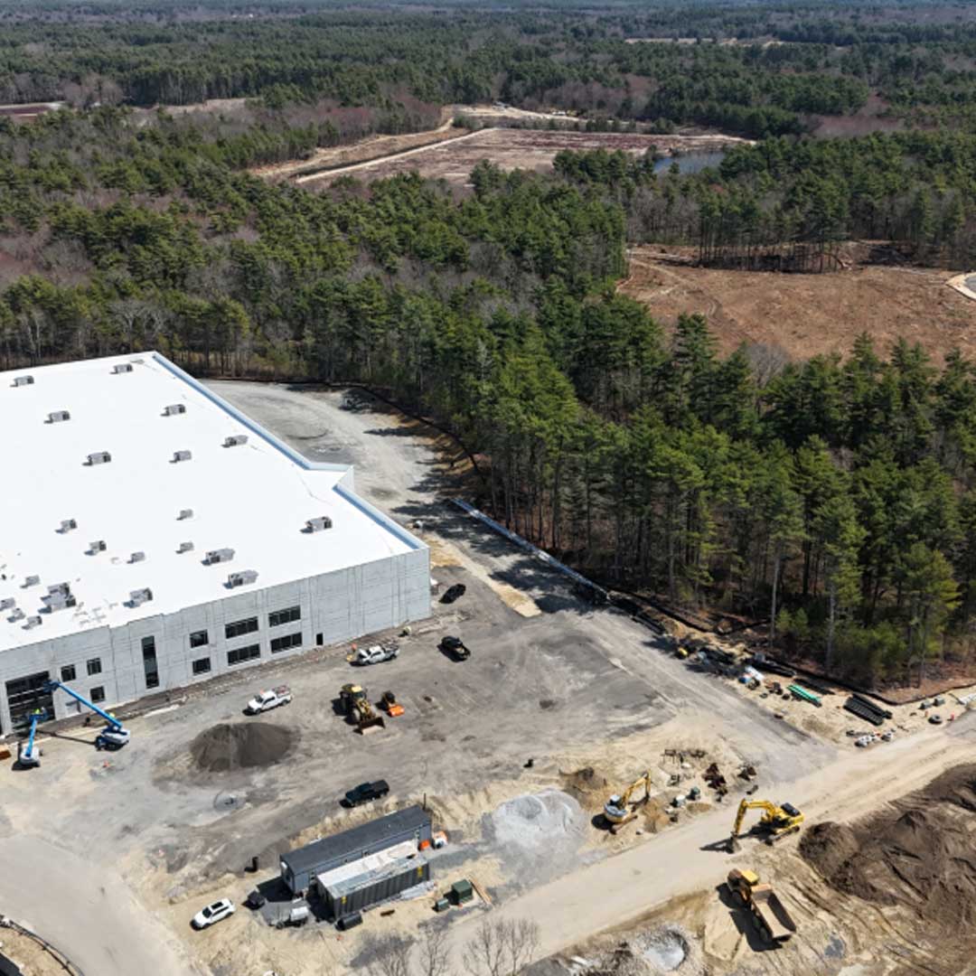 Aerial view of large industrial warehouse under construction with white roof, heavy equipment, and forested surroundings.