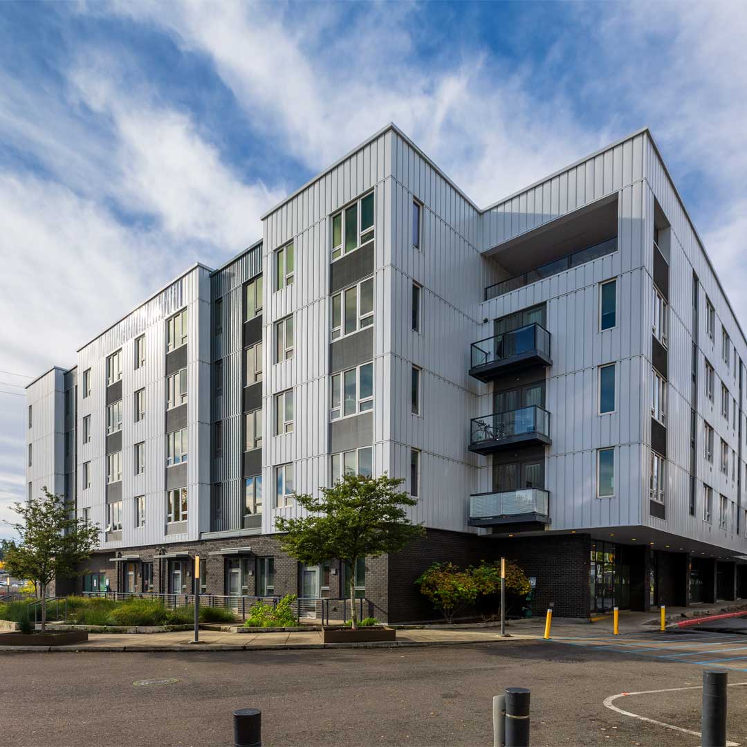 Modern multi-story apartment building with gray metal siding, balconies, large windows, and street-level entrances under a cloudy sky.
