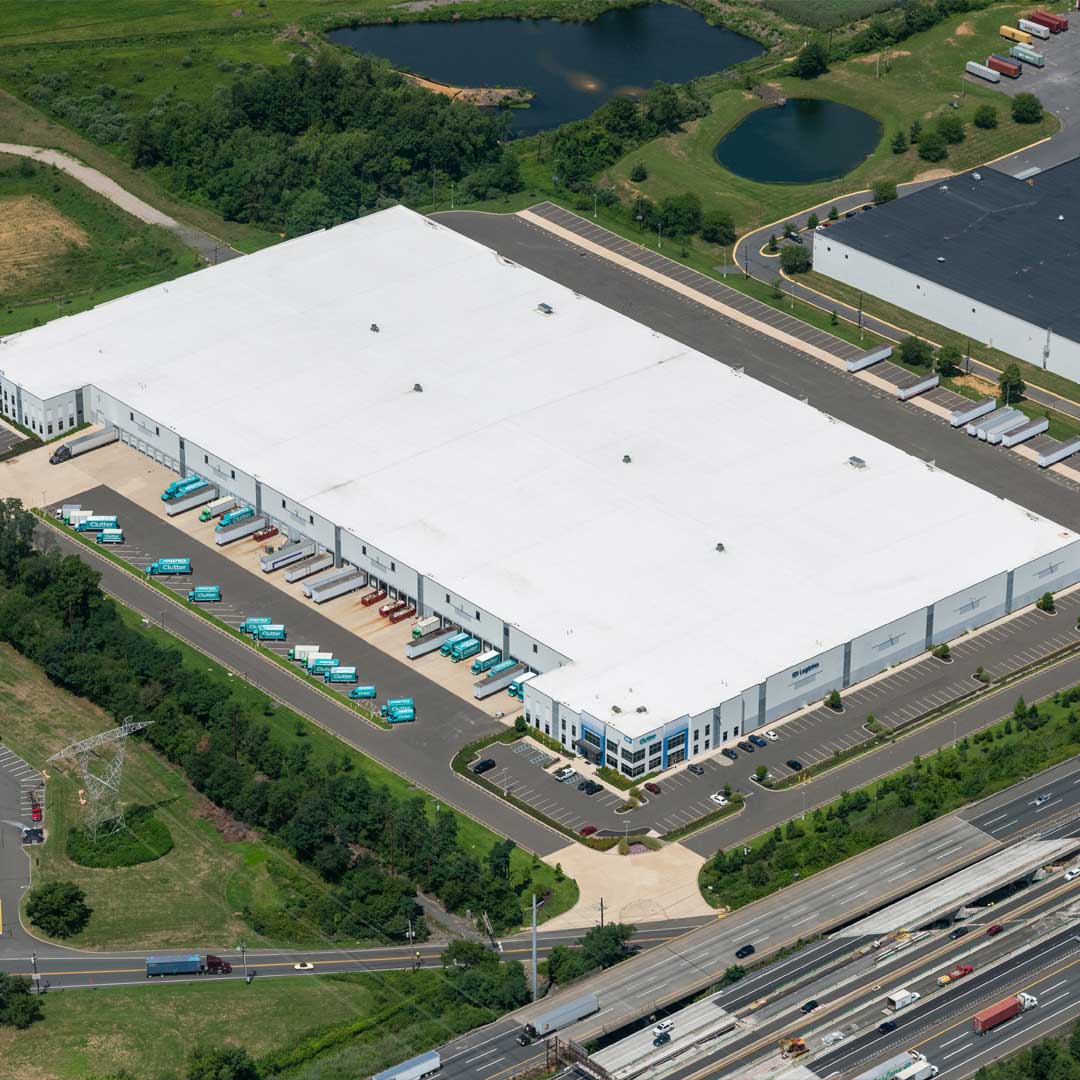 Aerial view of a large distribution warehouse with loading docks, delivery trucks, parking areas, and highway access in an industrial area.