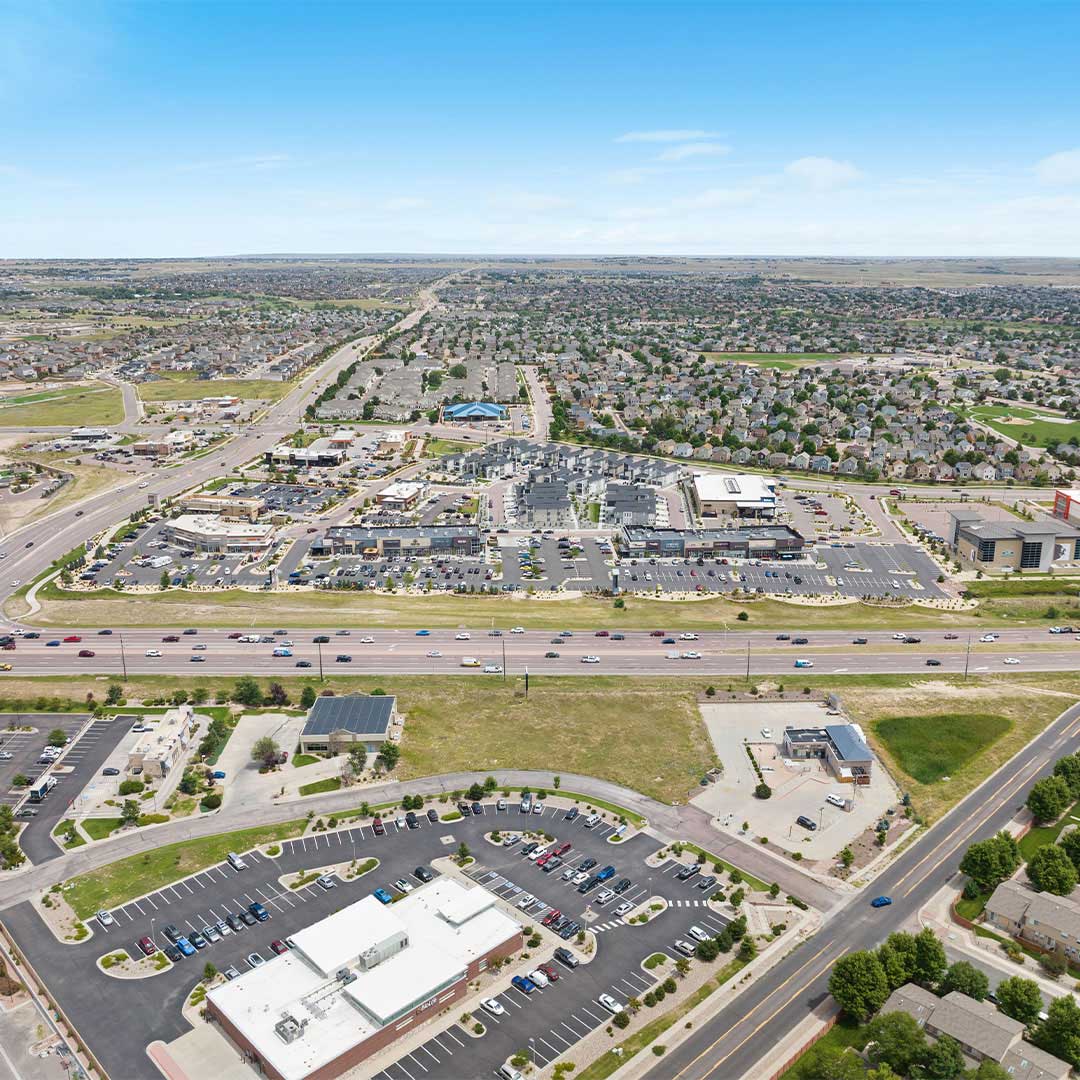 Aerial view of an unanchored strip shopping center with retail stores, parking lots, and highway access in a suburban neighborhood.