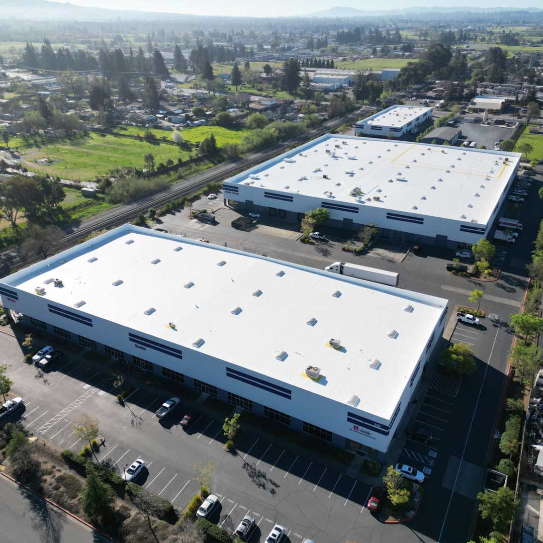 Aerial view of modern industrial warehouse complex with large white roofs, parking areas, and surrounding green landscape.