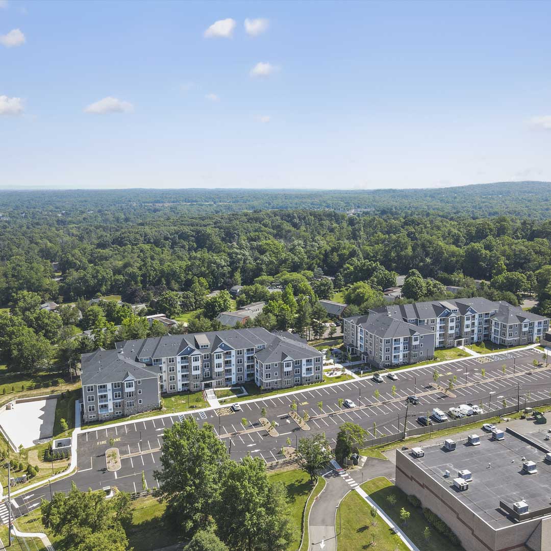 Aerial view of a modern apartment community with multiple residential buildings, parking lots, and surrounding green landscape in a suburban area.