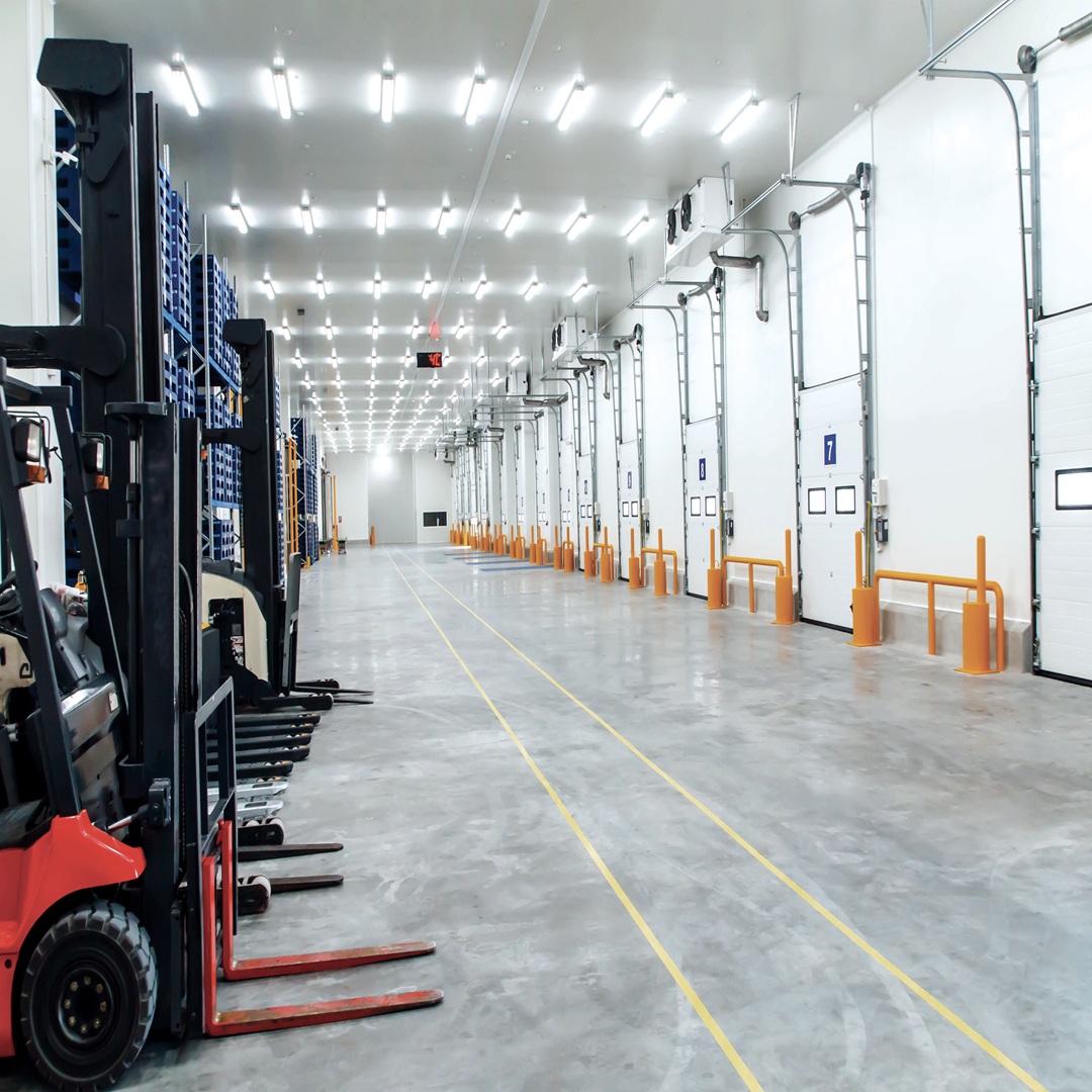Modern industrial warehouse interior with multiple loading dock doors, forklifts, pallet racking, and polished concrete floors in a distribution center.