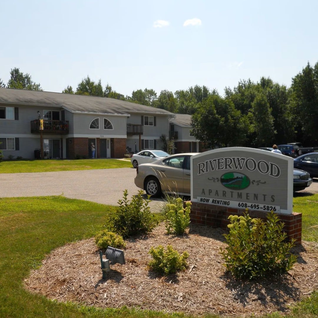 Exterior view of Riverwood Apartments community with landscaped entrance sign, residential buildings, and parking area.