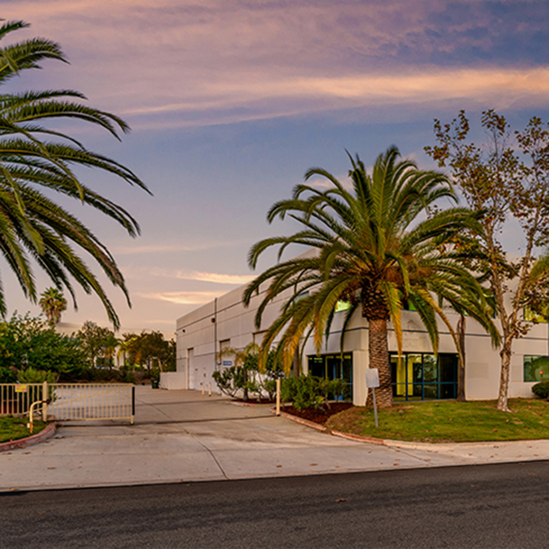 Exterior of industrial warehouse facility in Escondido, CA at dusk, featuring palm trees, gated driveway, and modern tilt-up construction.