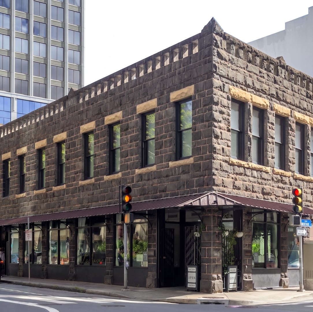 Historic 1902 two-story rusticated stone corner building with retail storefronts, black-framed windows, and metal awning in urban downtown.