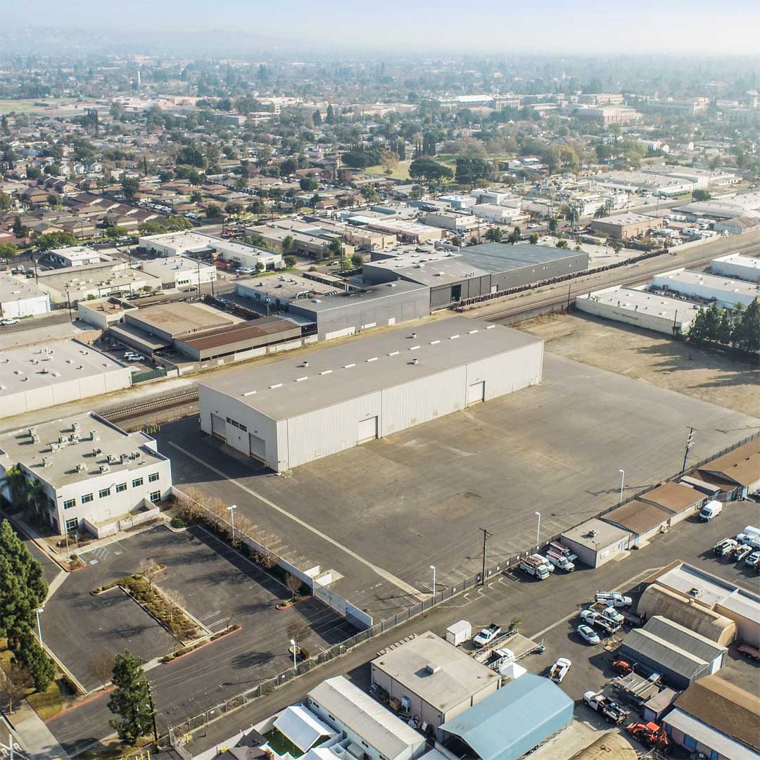 Drone view of an industrial warehouse with a large paved yard, surrounded by commercial buildings in Orange, California.
