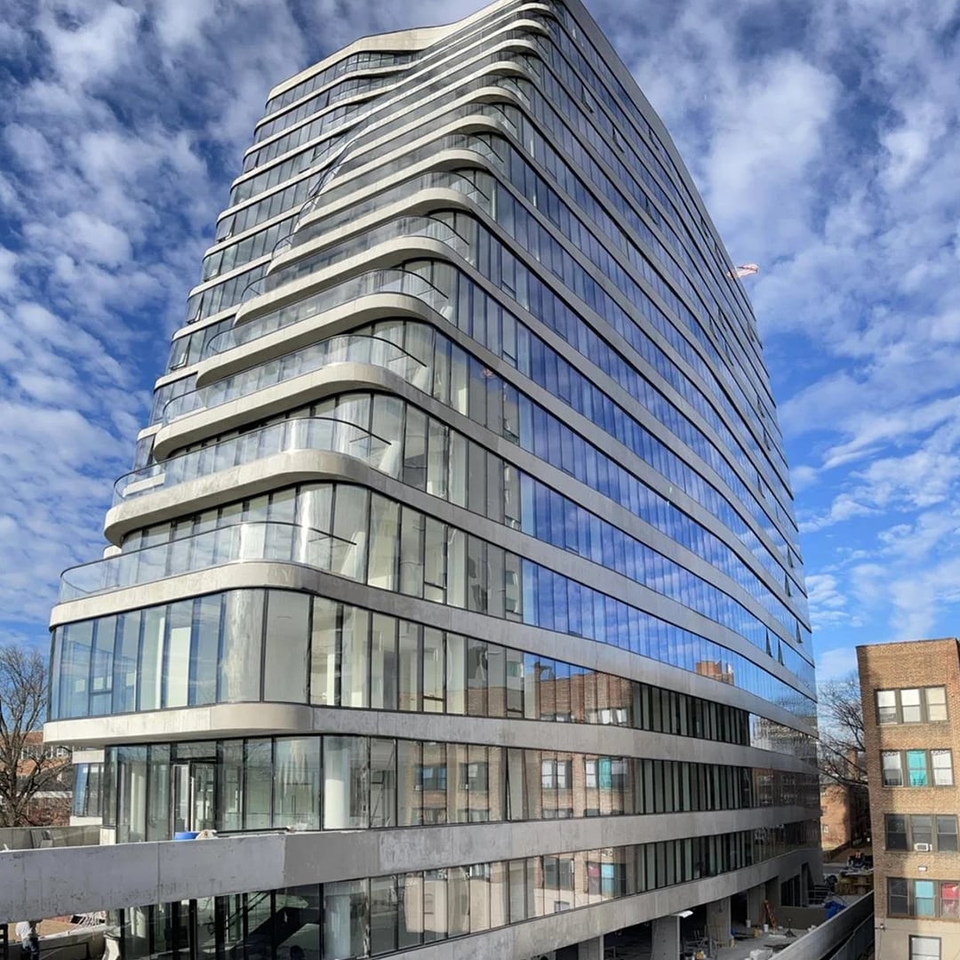 Exterior of a modern glass high-rise with curved balconies in an urban commercial district under a blue sky.