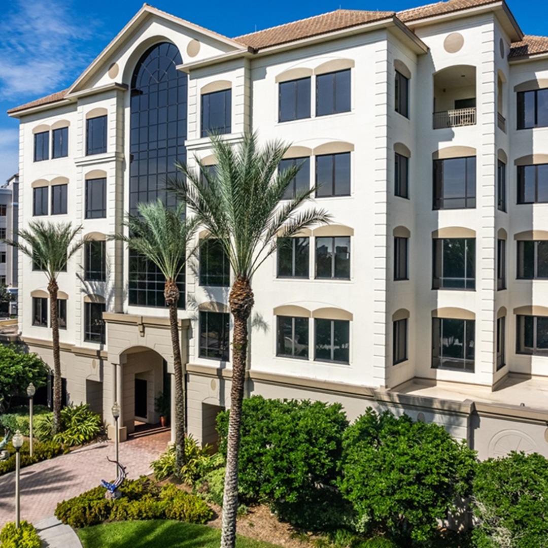 Exterior view of a modern multi‑story office building surrounded by palm trees and landscaped greenery in a bright, sunny setting.