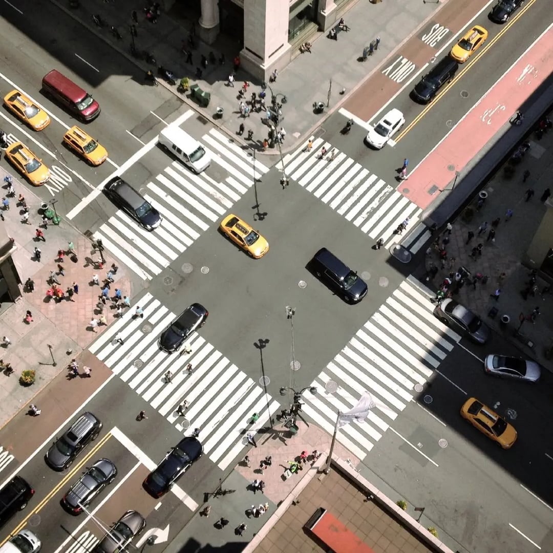 Overhead view of a busy city intersection with crosswalks, cars, taxis, and pedestrians moving through the streets.