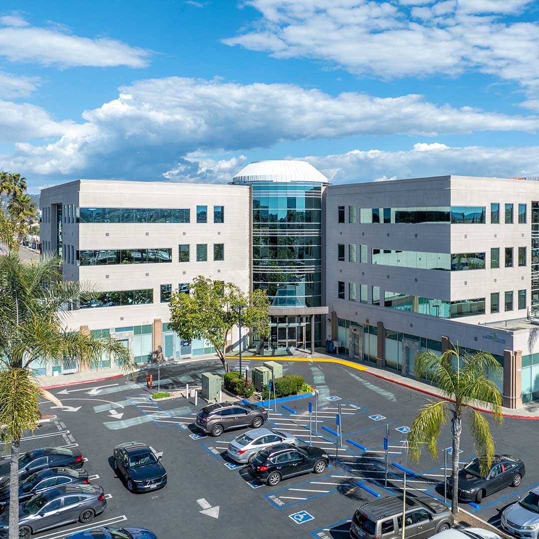 Modern three‑story office building with glass entrance, palm trees, and a busy parking lot under a partly cloudy sky.