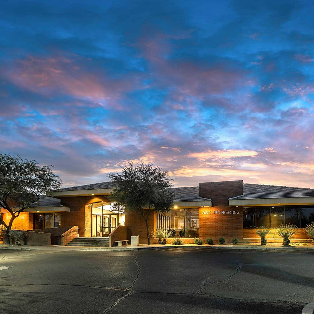 Brick office building at dusk with warm lights glowing, framed by desert plants under a colorful blue and pink sky.