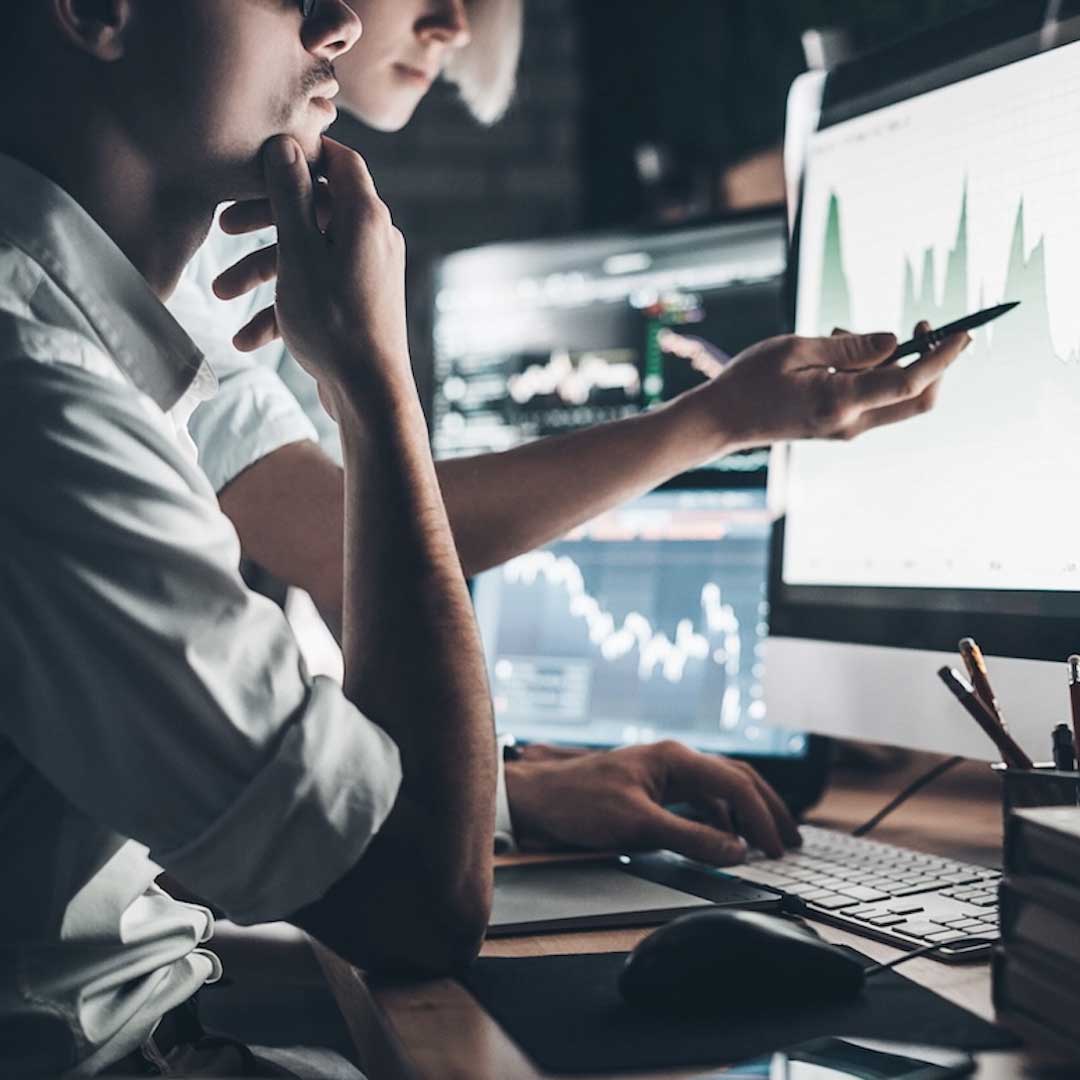 Team examining financial analytics on computer screens, pointing at charts showing market movements and performance trends.
