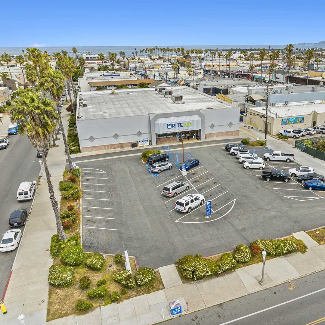 Aerial view of a Rite Aid store with parked cars, palm trees along the street, and the ocean visible behind the coastal neighborhood.
