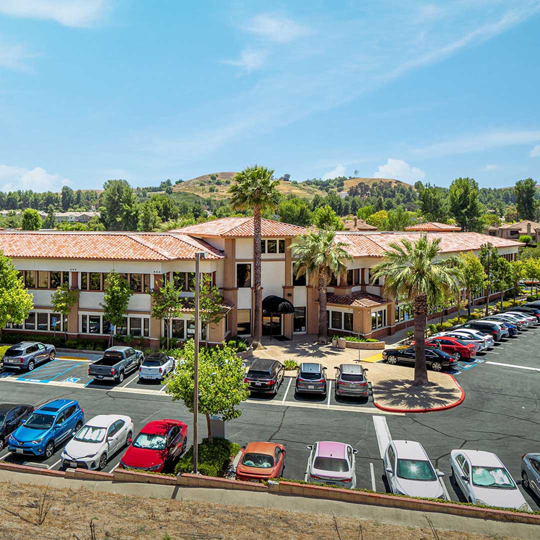 Two‑story office building with red‑tile roof, palm trees, and a busy parking lot set against green hills under a bright blue sky.