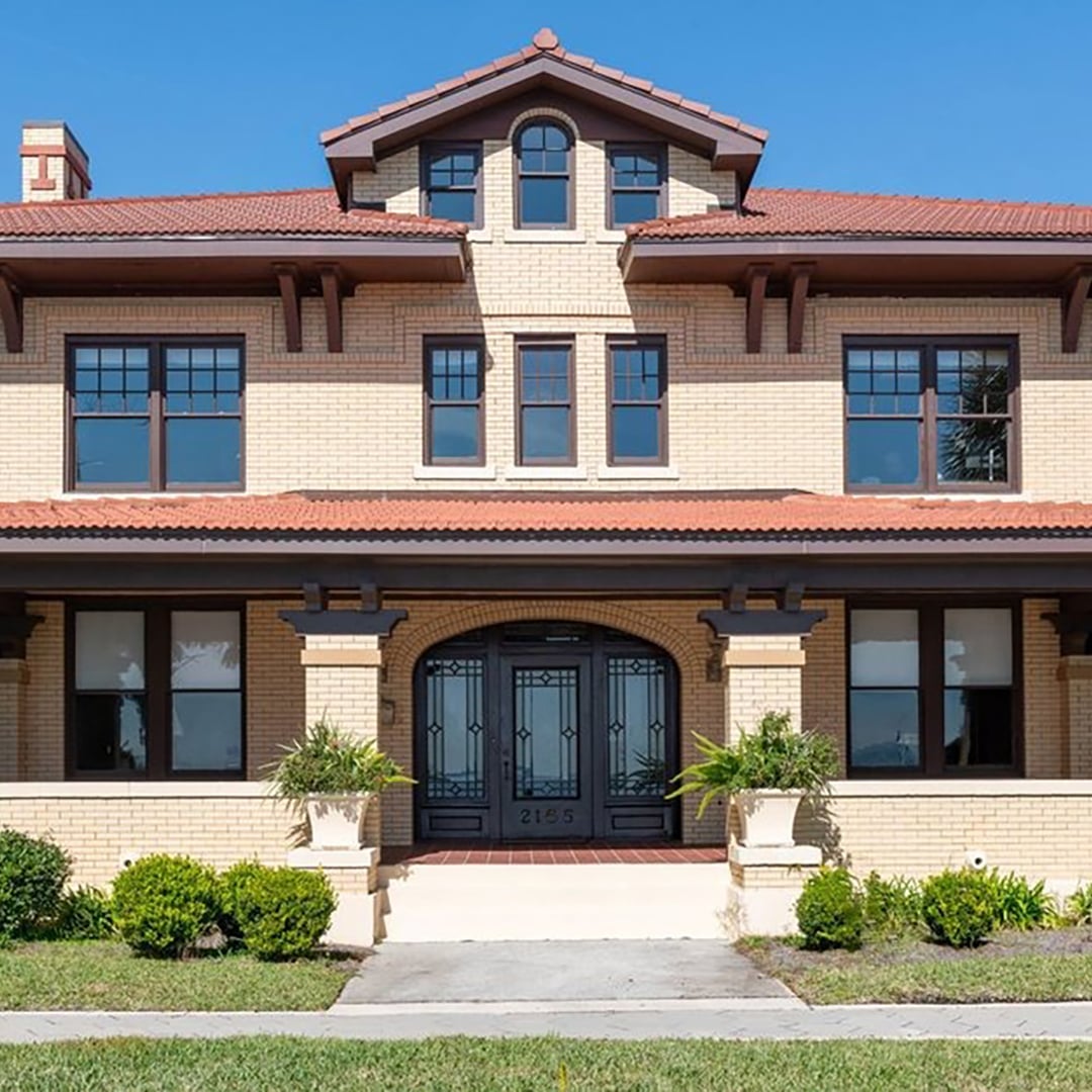 Front elevation of a historic two-story brick home with large windows, red tile roof, and landscaped entry walkway.