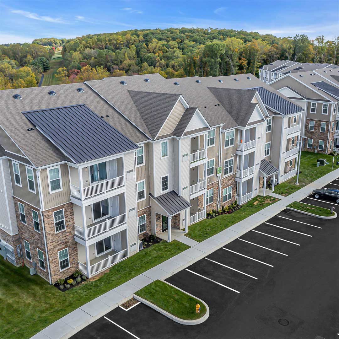 Aerial view of Mi Place, a modern 3-story apartment community with balconies, stone accents, and parking lot, set against a wooded hillside.